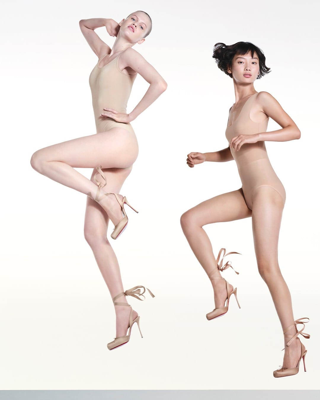 Two models wearing nude bodysuits and lace-up high heels posing dynamically against a clean white seamless background.