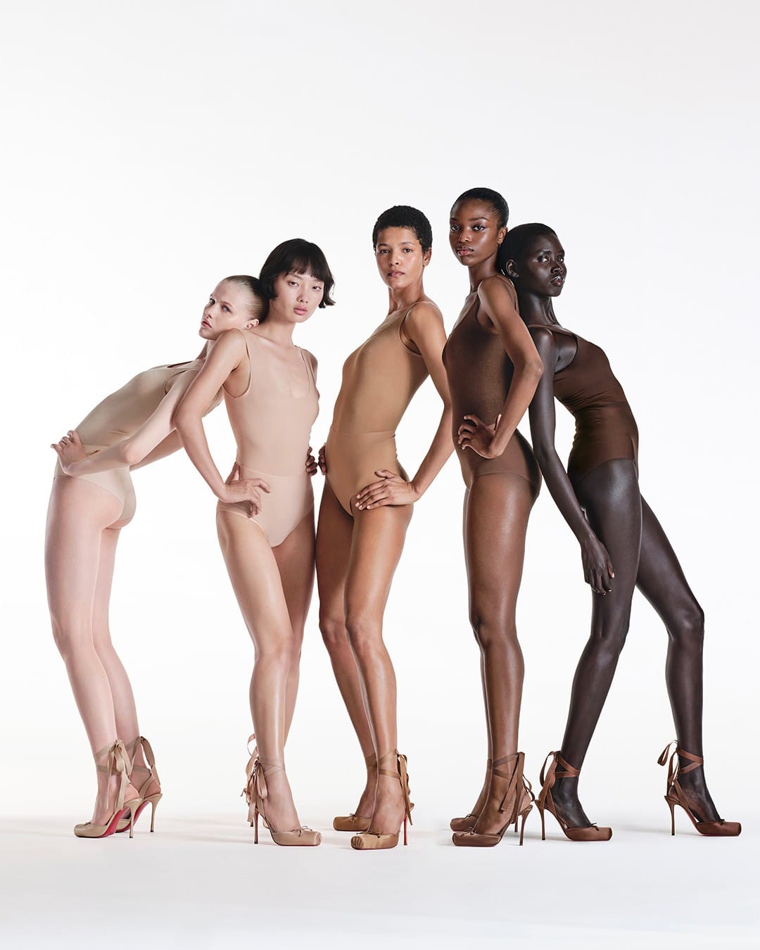 Five models posing in bodysuits and lace-up high heels against a white studio seamless backdrop for Christian Louboutin.