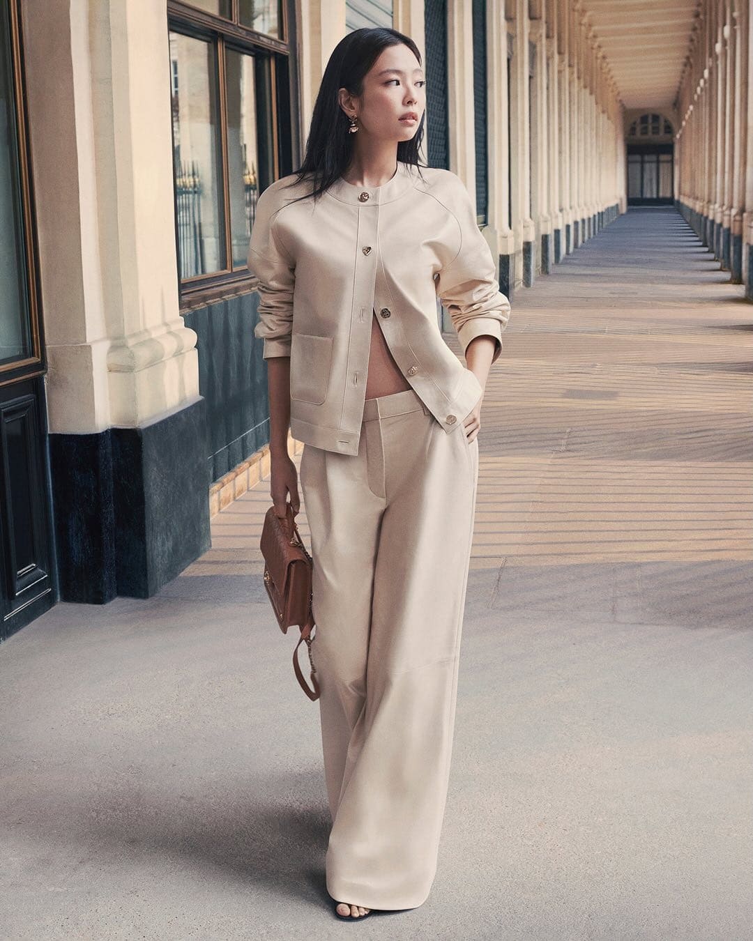 Model Jennie wearing a neutral beige jacket and matching trousers holding a small brown handbag along an arcade corridor in Paris.