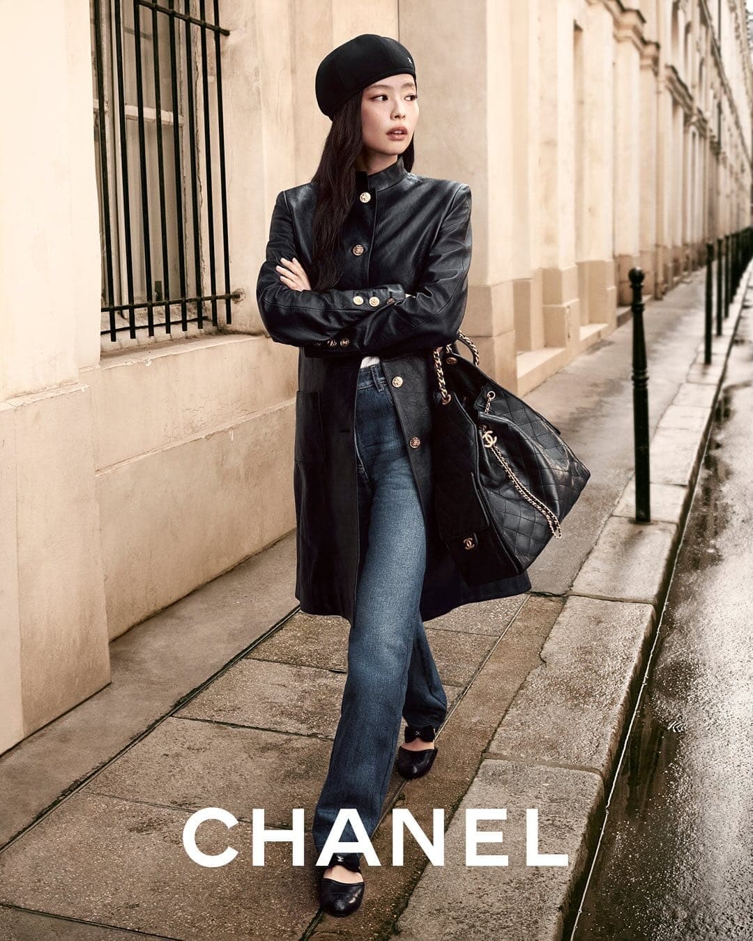 Model Jennie wearing a black beret, leather coat, and denim jeans while standing on a wet stone street by historic building facades.