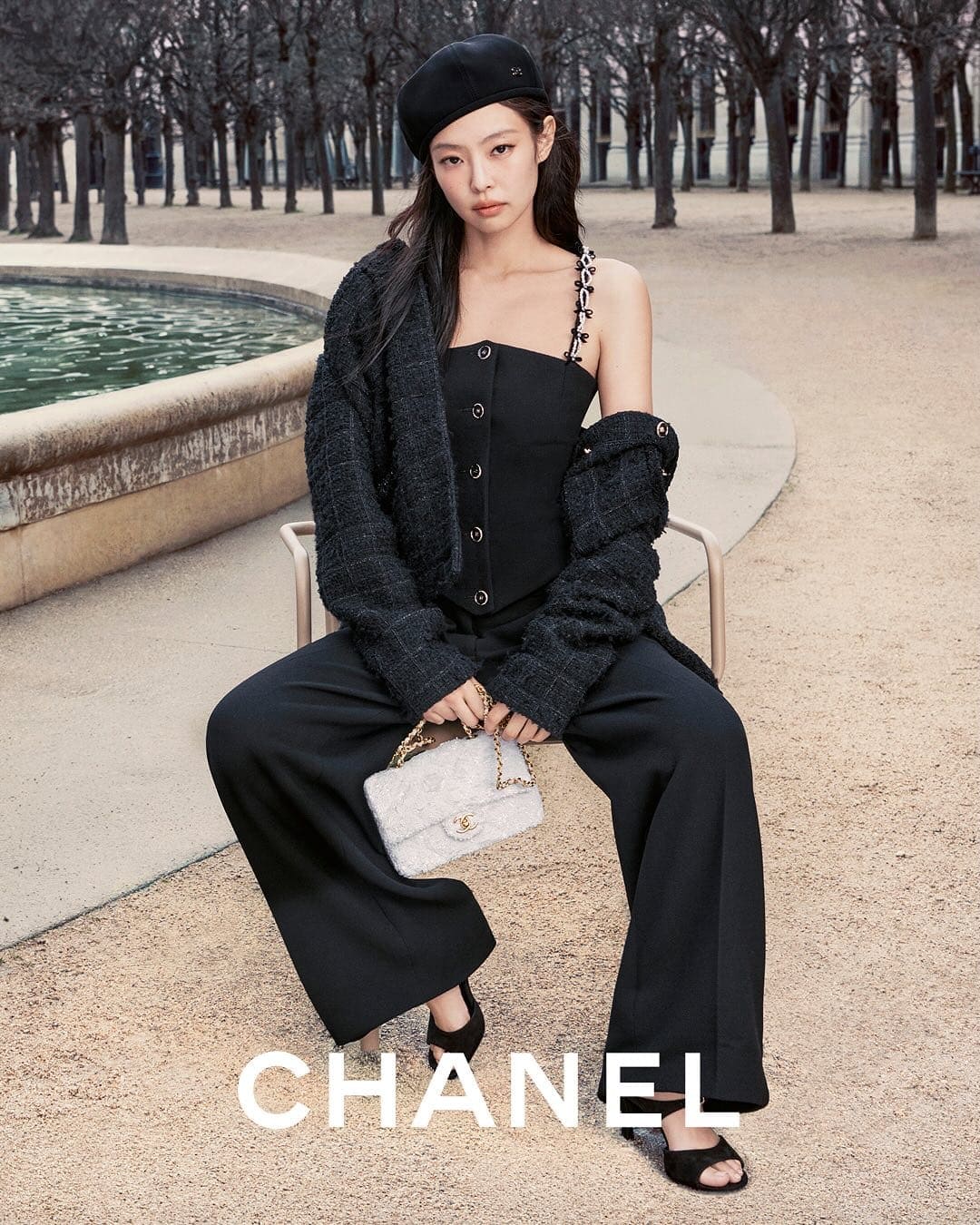 Model Jennie wearing a black beret, tweed jacket, top, and wide trousers, holding a white handbag outdoors near a fountain.