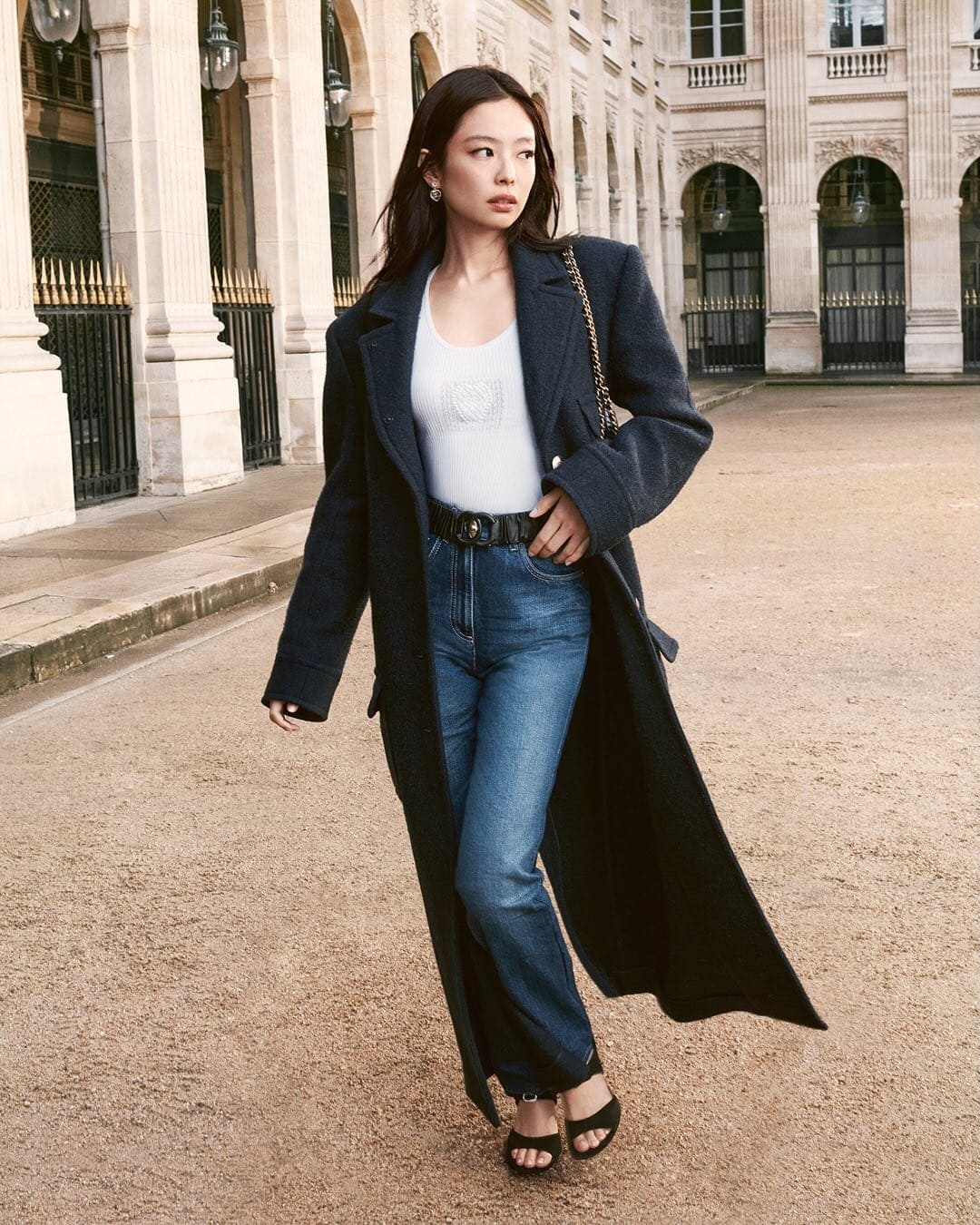 Jennie wearing a dark wool coat and blue jeans standing outdoors near classical arcade architecture.