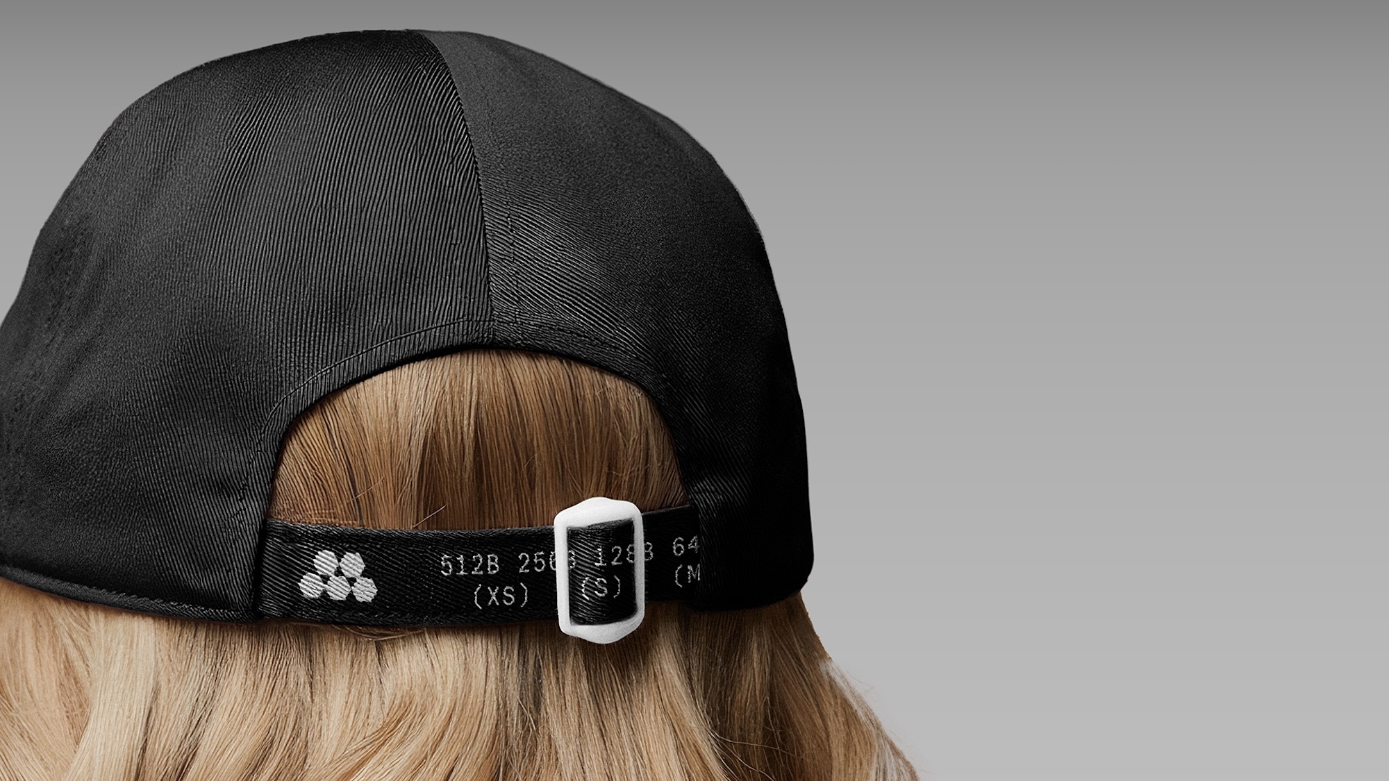 Rear view of a black cap with a white adjustable strap on a blonde person against a neutral grey background