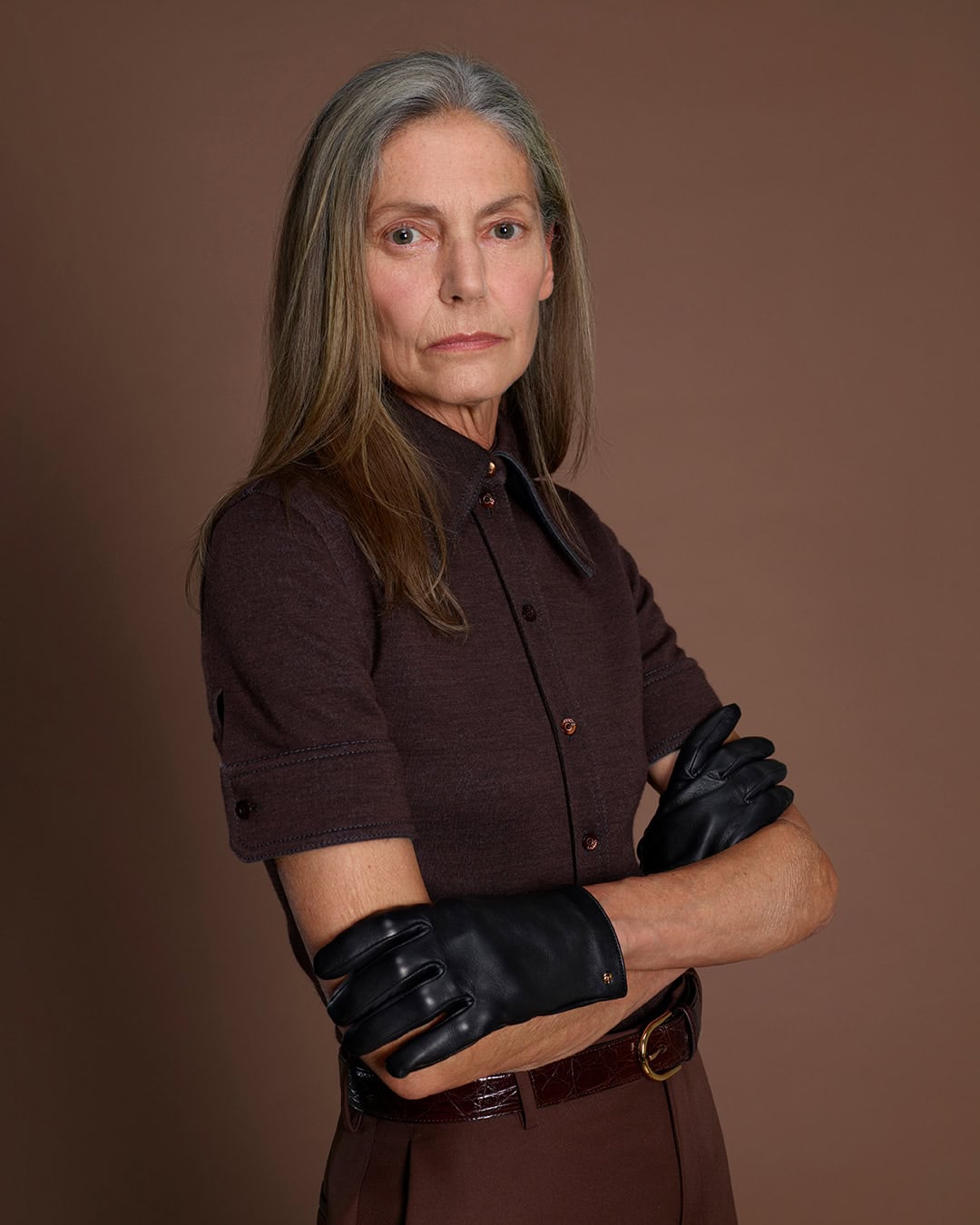 Elderly woman with long grey hair wearing a brown short-sleeve buttoned shirt and black leather gloves against a brown background.