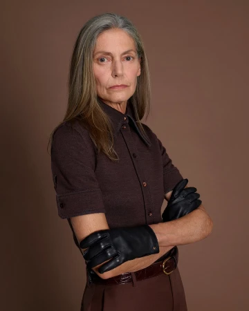 Elderly woman with long grey hair wearing a brown short-sleeve buttoned shirt and black leather gloves against a brown background.