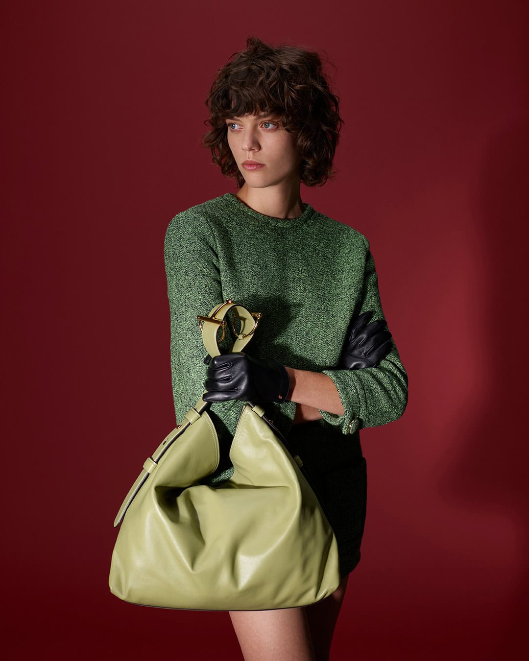 Model in green sweater and leather gloves holding a light green handbag against a deep red background for Gucci.