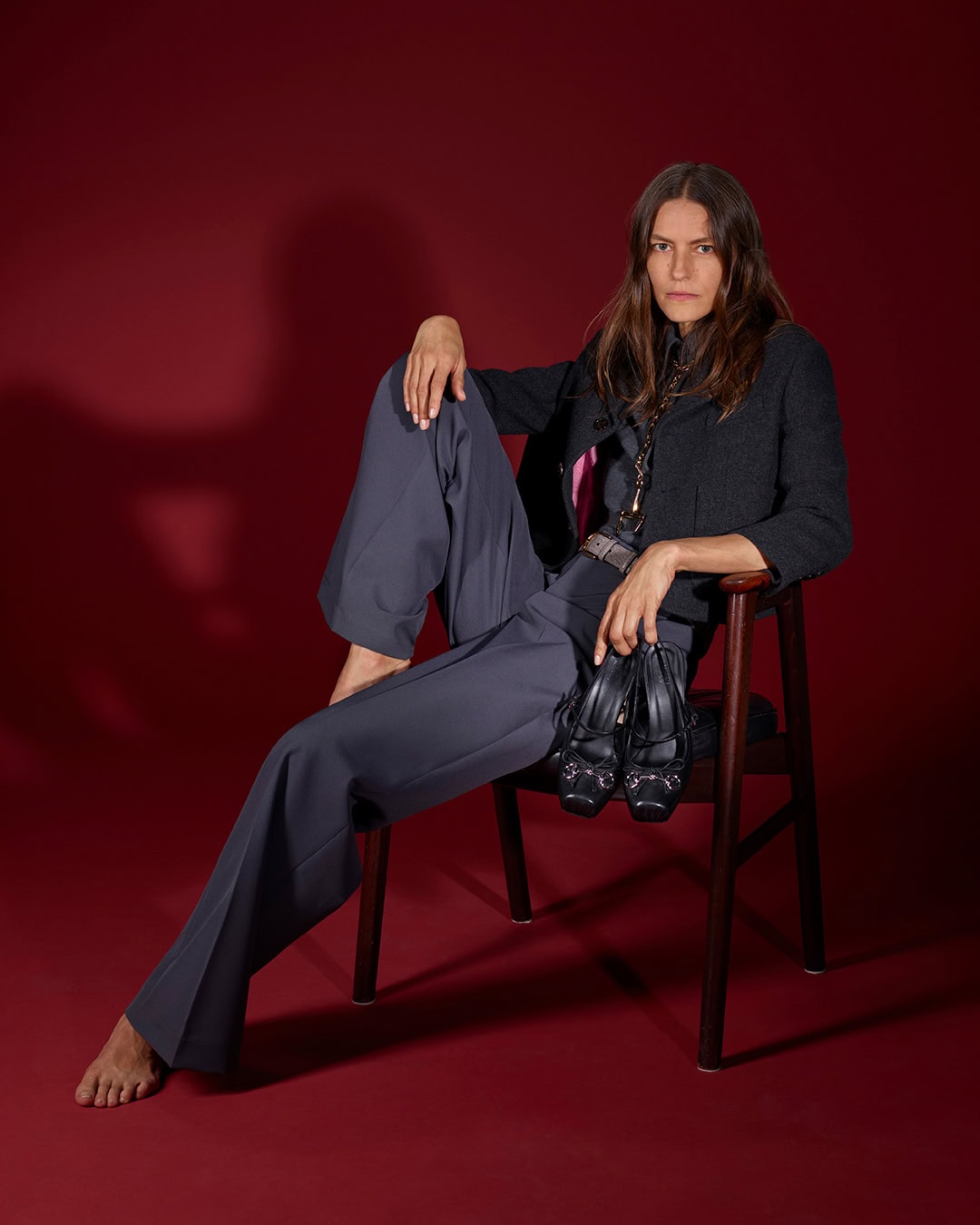 Model seated on a dark wooden chair against a deep red backdrop, holding black shoes and wearing grey trousers and a dark jacket.