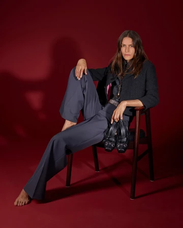 Model seated on a dark wooden chair against a deep red backdrop, holding black shoes and wearing grey trousers and a dark jacket.
