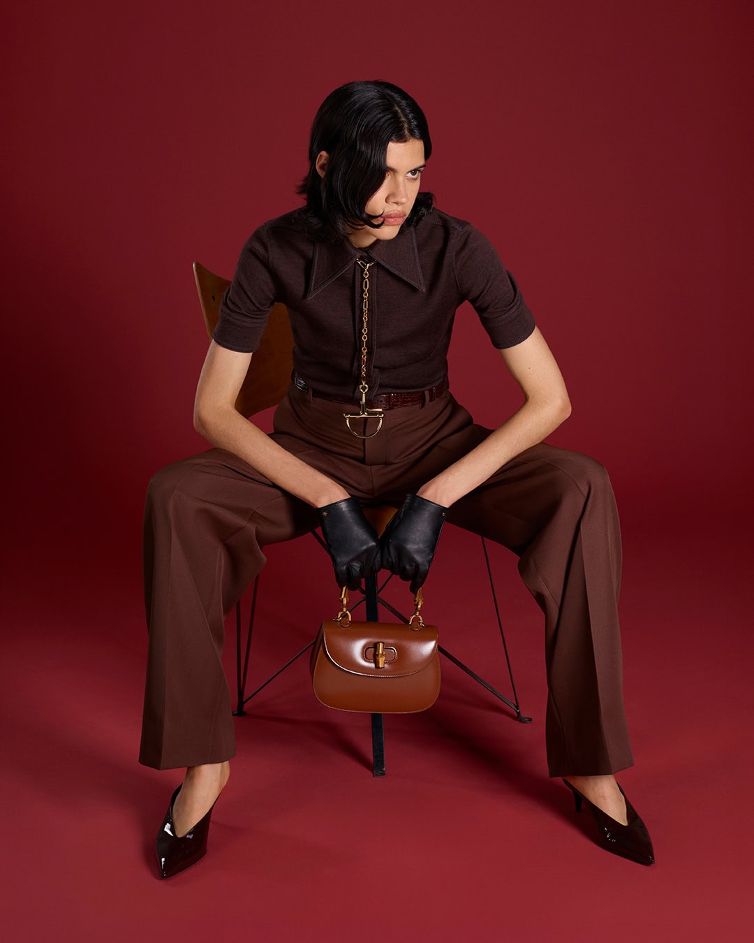 Model seated on a chair wearing a brown polo shirt and trousers, holding a brown leather handbag.