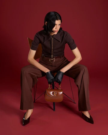 Model seated on a chair wearing a brown polo shirt and trousers, holding a brown leather handbag.