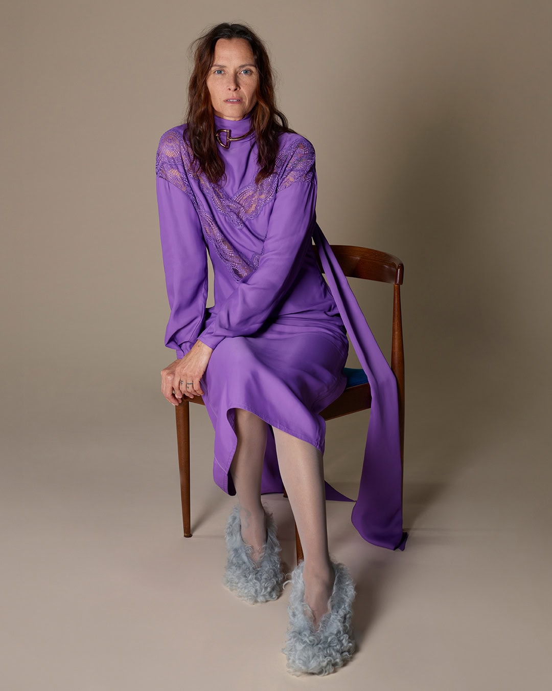 Model seated on a wooden chair wearing a purple dress and textured fluffy blue shoes against a plain neutral background.