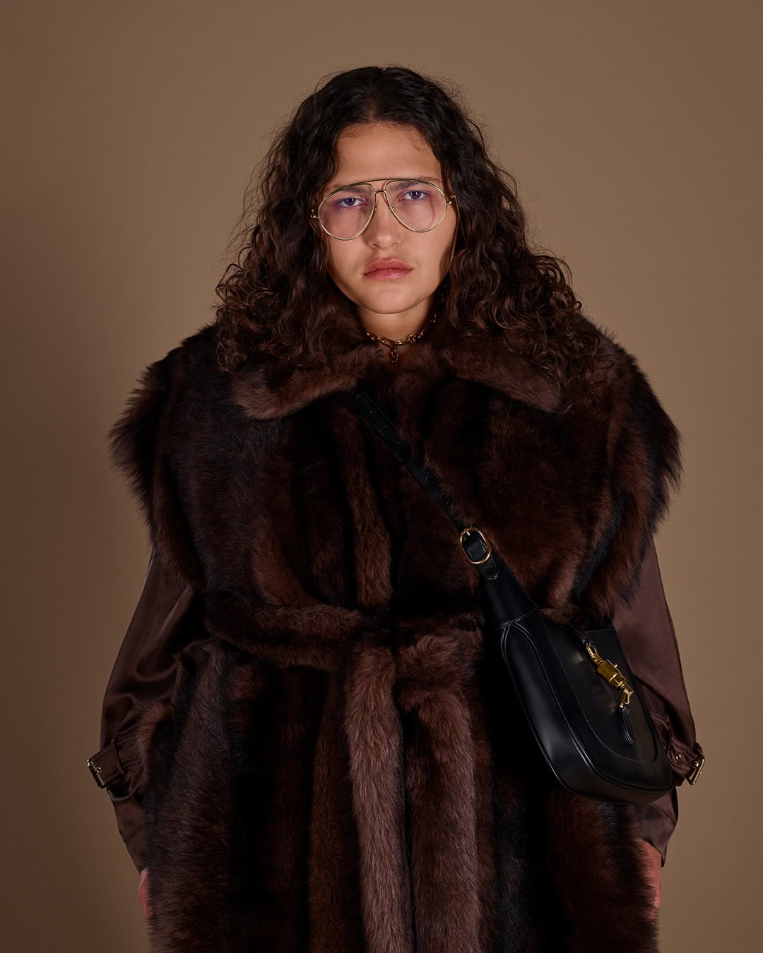 Model wearing large glasses and a dark brown fur coat carrying a black leather shoulder bag against a neutral brown backdrop