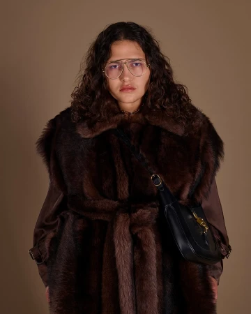 Model wearing large glasses and a dark brown fur coat carrying a black leather shoulder bag against a neutral brown backdrop