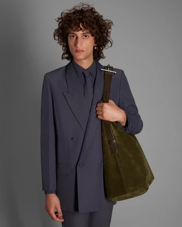 Man wearing a slate blue suit holding an olive green shoulder bag against a grey background