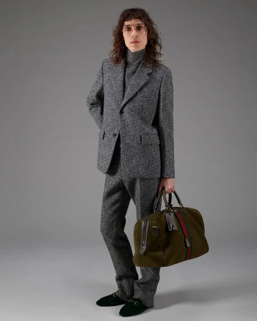Model in grey tweed suit and turtleneck wearing glasses, holding a green duffel bag against a grey background