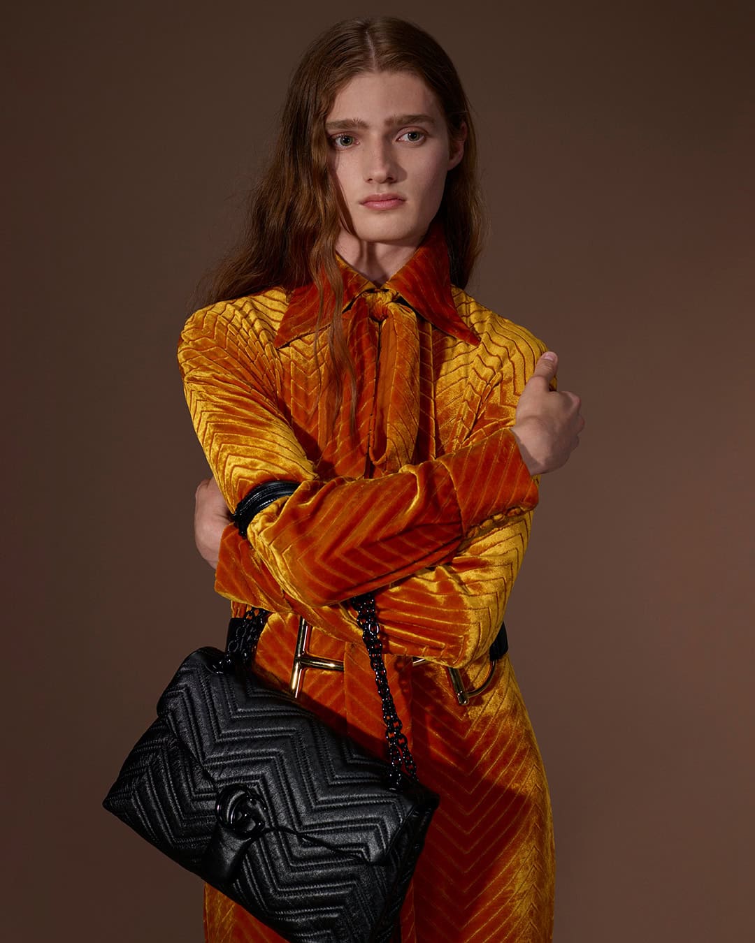 Model in orange textured coat holding a black quilted handbag against a warm brown background