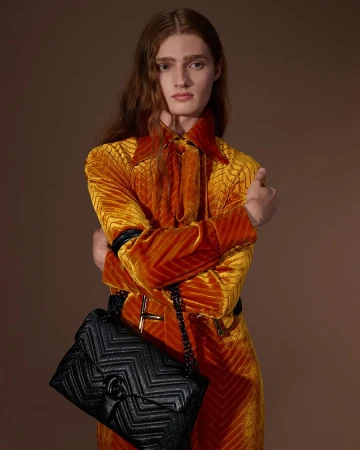 Model in orange textured coat holding a black quilted handbag against a warm brown background