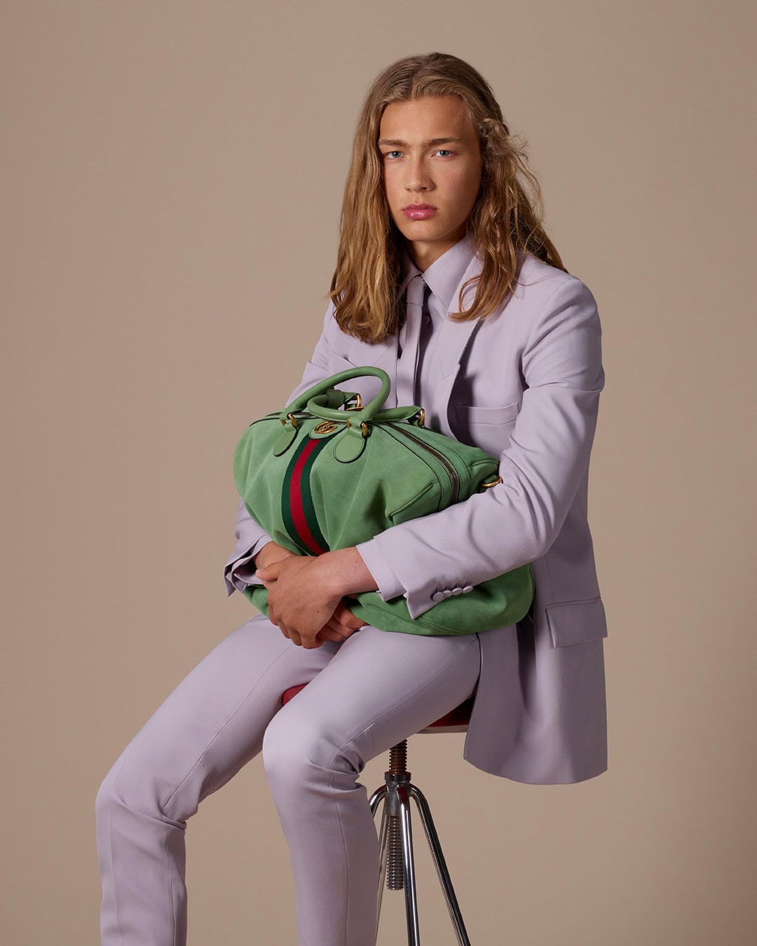 Model with long blonde hair seated on a stool holding a green duffel bag, wearing a lavender suit jacket and trousers against a neutral backdrop.