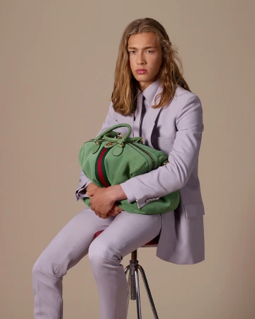 Model with long blonde hair seated on a stool holding a green duffel bag, wearing a lavender suit jacket and trousers against a neutral backdrop.