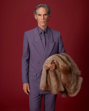 Man wearing a purple suit holding a furry brown coat against a dark red studio seamless background