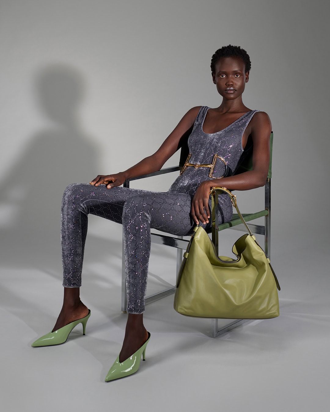 Black woman seated on a chrome chair in a grey jumpsuit holding an olive green handbag for Gucci Fall 2025 campaign.