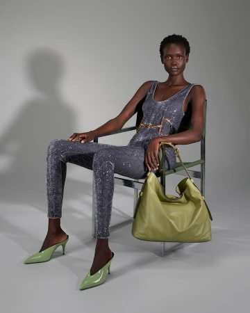 Black woman seated on a chrome chair in a grey jumpsuit holding an olive green handbag for Gucci Fall 2025 campaign.