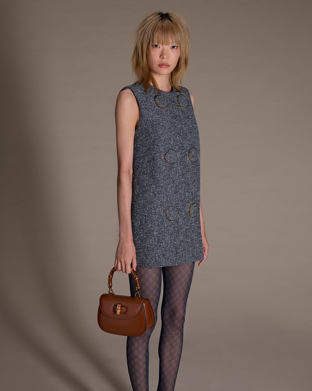 Model in grey tweed dress wearing patterned tights holding a brown handbag against grey seamless backdrop
