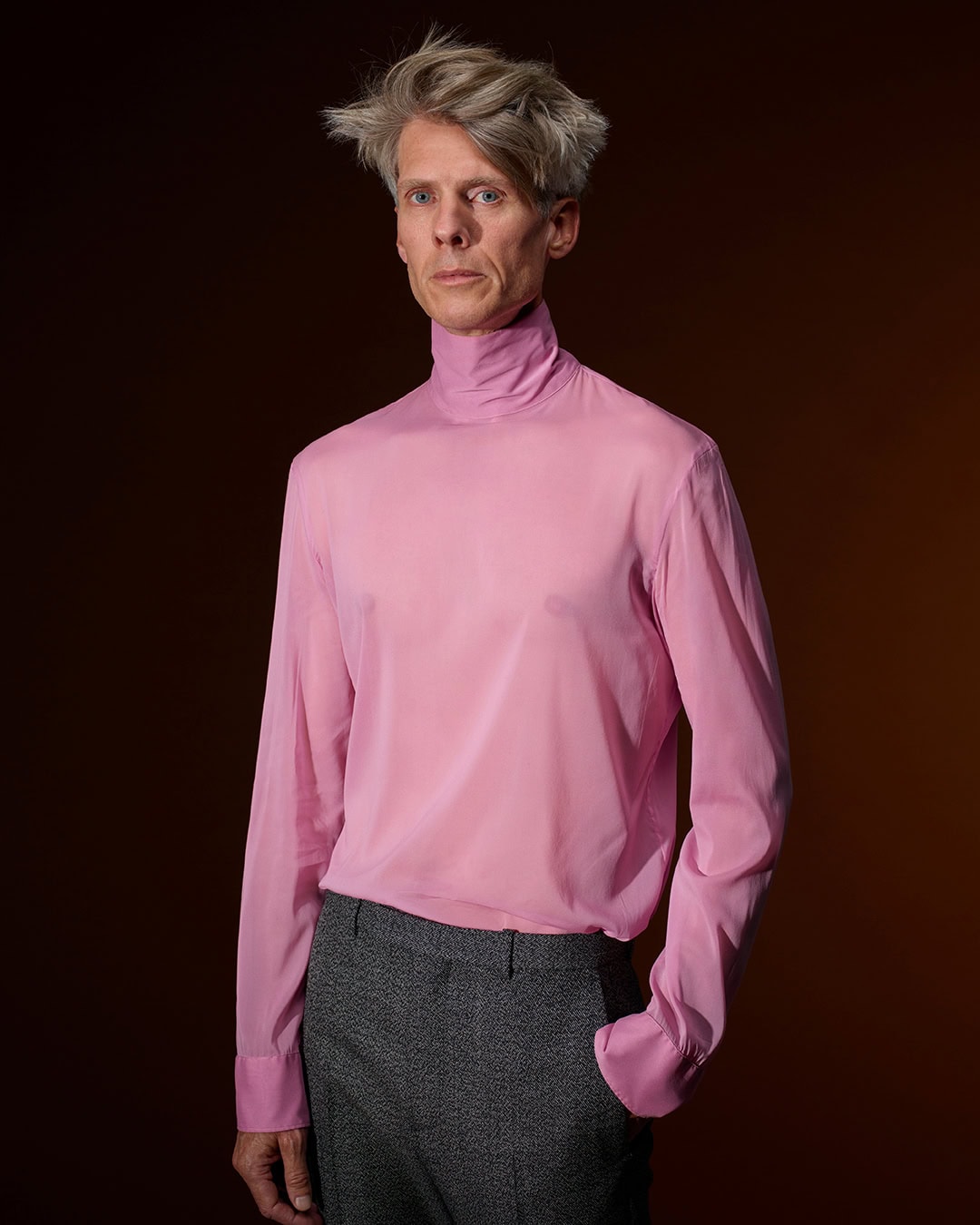 Man wearing a sheer pink turtleneck shirt and dark grey trousers against a dark gradient backdrop