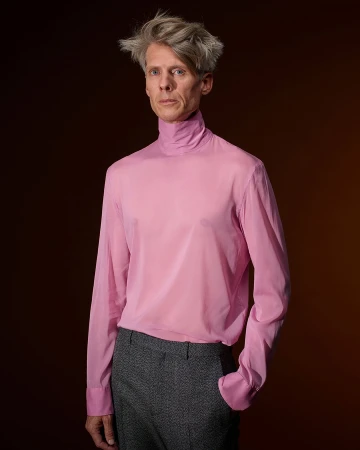 Man wearing a sheer pink turtleneck shirt and dark grey trousers against a dark gradient backdrop