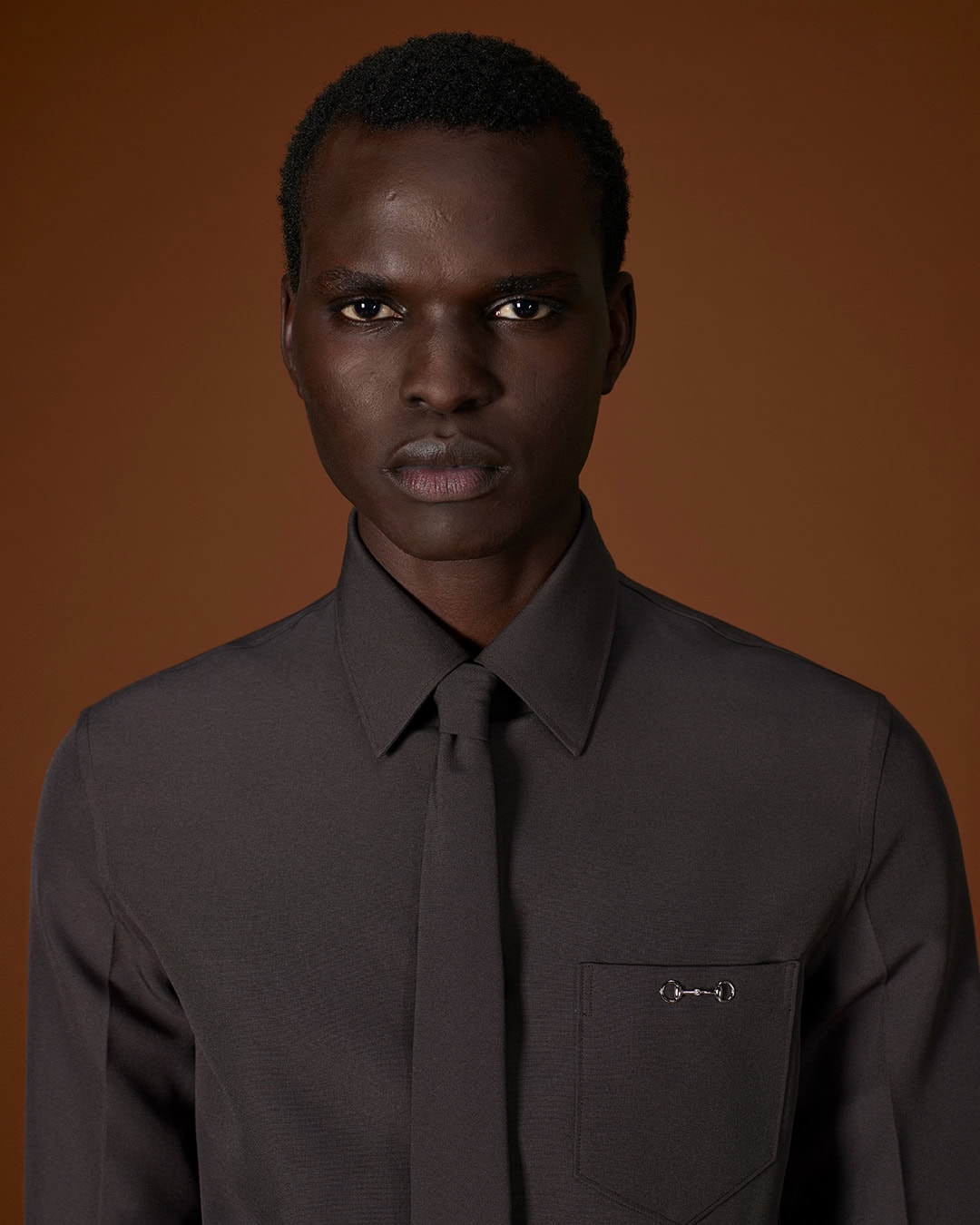 Portrait of a Black man in a dark grey button-down shirt and matching tie against a warm brown background for Gucci Fall 2025.
