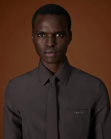 Portrait of a Black man in a dark grey button-down shirt and matching tie against a warm brown background for Gucci Fall 2025.