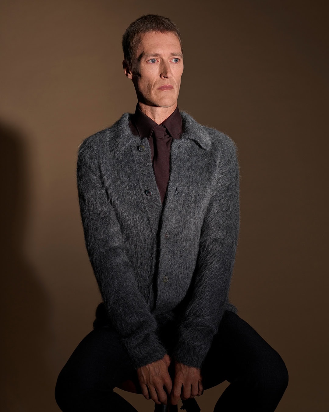 Man wearing a textured grey cardigan and dark shirt seated against a warm brown studio background.