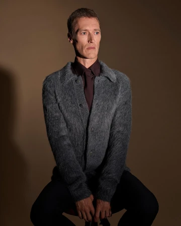 Man wearing a textured grey cardigan and dark shirt seated against a warm brown studio background.