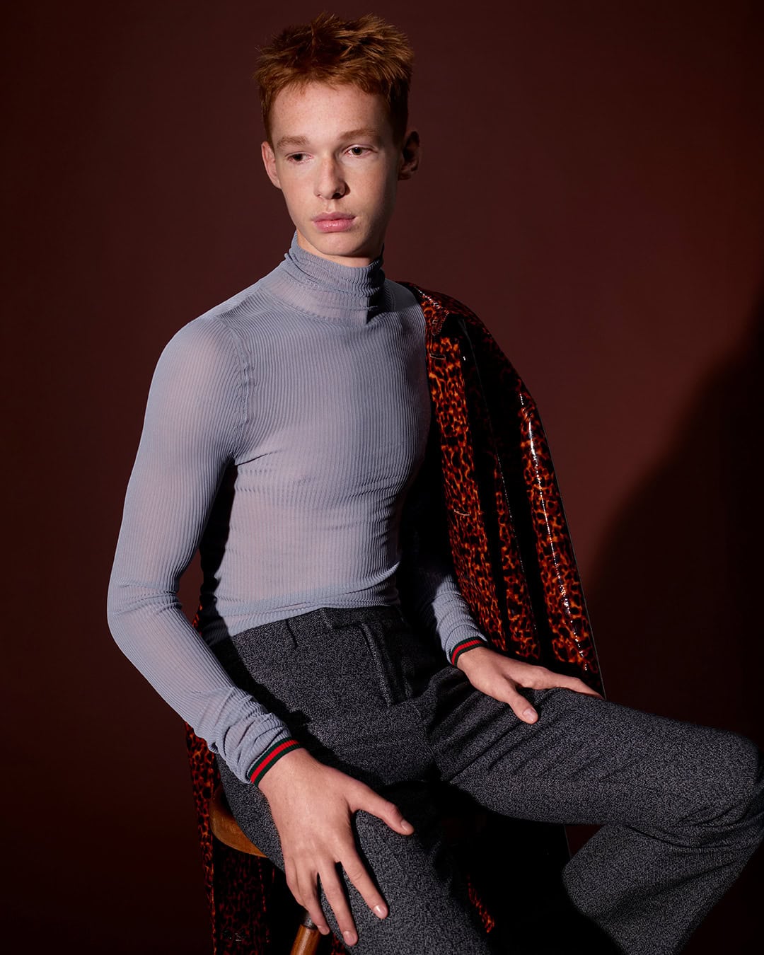 Young male model with red hair seated on a stool wearing a sheer grey turtleneck and dark grey trousers against a dark brown background.