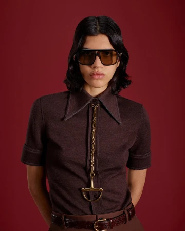 Model wearing oversized sunglasses and a brown button-up shirt against a deep red studio backdrop