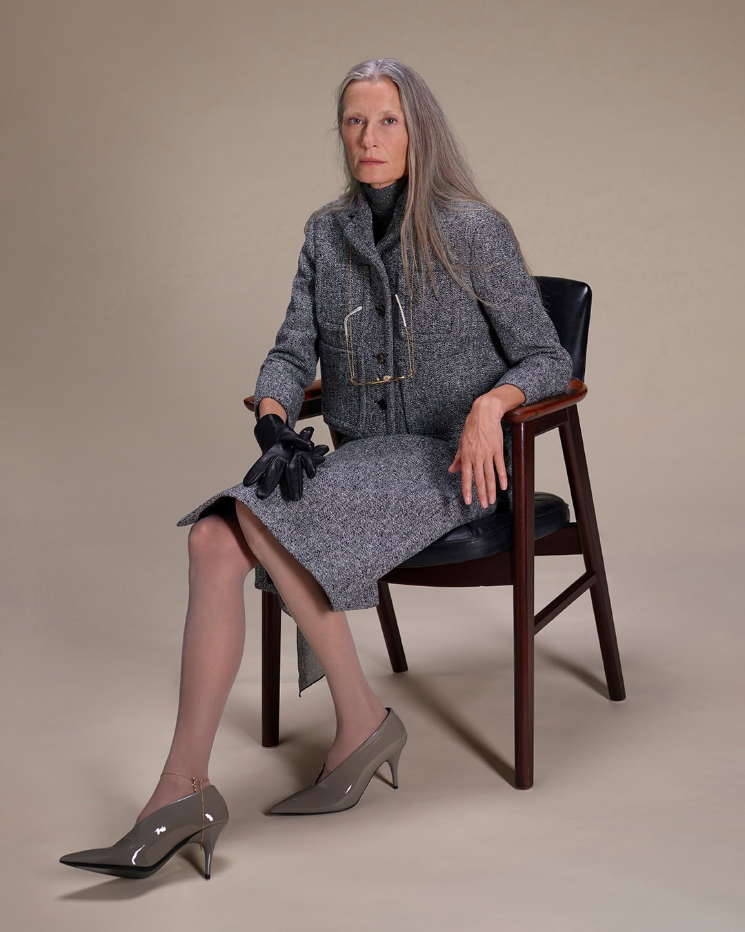 Elderly woman with long grey hair wearing a grey tweed skirt suit and heels, seated in an armchair against a neutral background.