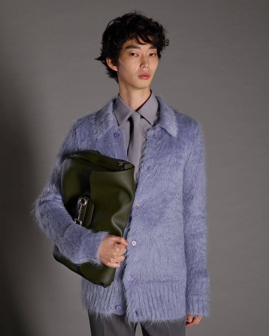 Man wearing a textured blue cardigan holding a dark green leather bag against a neutral grey background