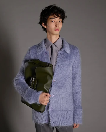 Man wearing a textured blue cardigan holding a dark green leather bag against a neutral grey background