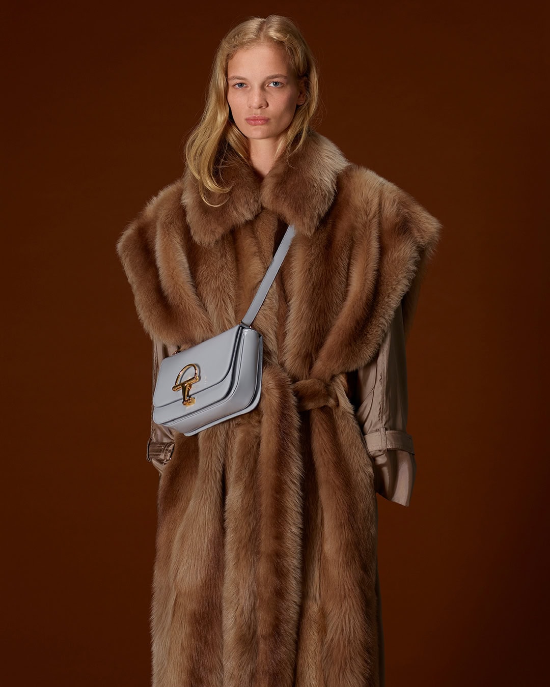 Model in brown fur coat wearing a light blue crossbody bag against a dark background for Gucci Fall 2025 Ad Campaign.