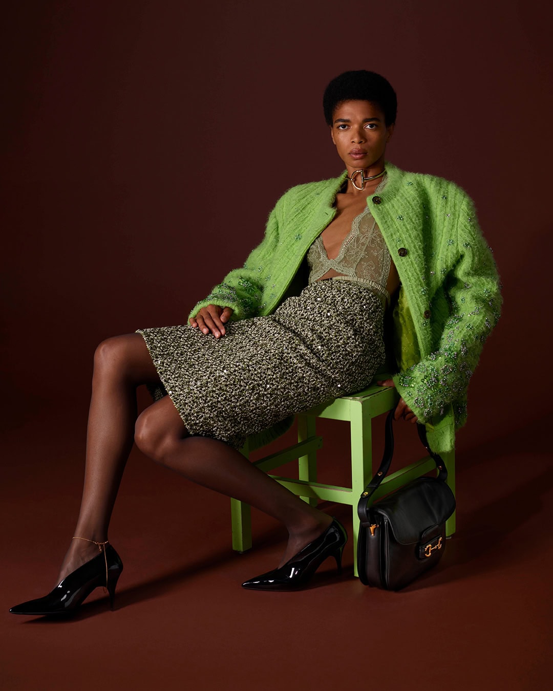 Model wearing a bright green textured jacket and patterned skirt seated on a matching green stool against a dark brown background.
