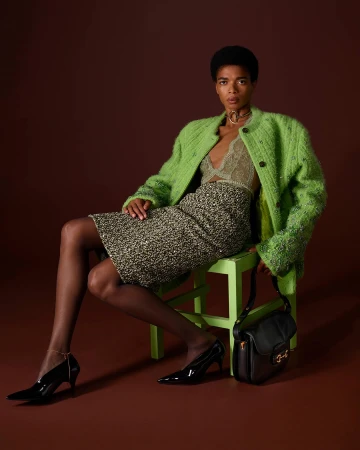 Model wearing a bright green textured jacket and patterned skirt seated on a matching green stool against a dark brown background.
