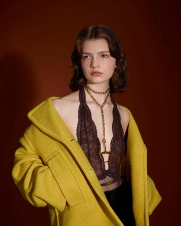 Model wearing a yellow coat and lace top with a gold chain necklace against a dark red backdrop