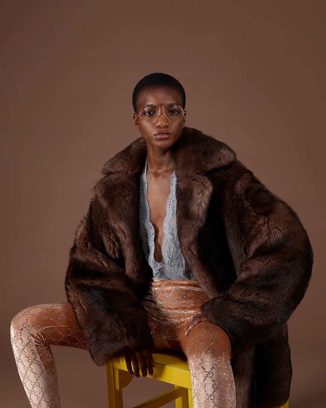 Model seated wearing a large brown fur coat over a grey lace top and patterned trousers against a brown backdrop