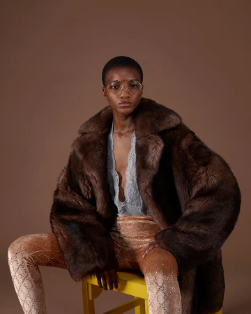 Model seated wearing a large brown fur coat over a grey lace top and patterned trousers against a brown backdrop