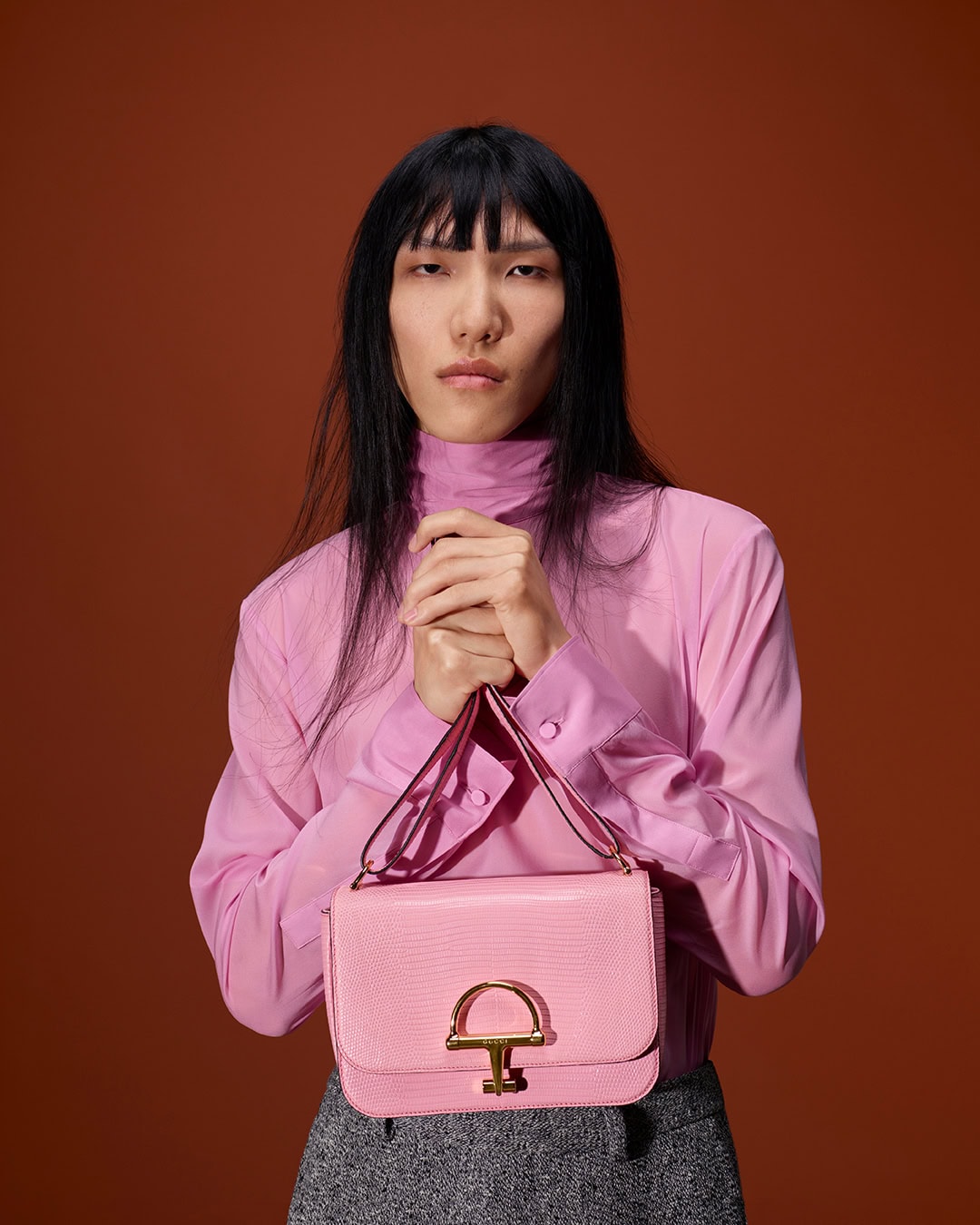 Model with long dark hair wearing a pink high-neck blouse holding a light pink handbag against a solid brown background