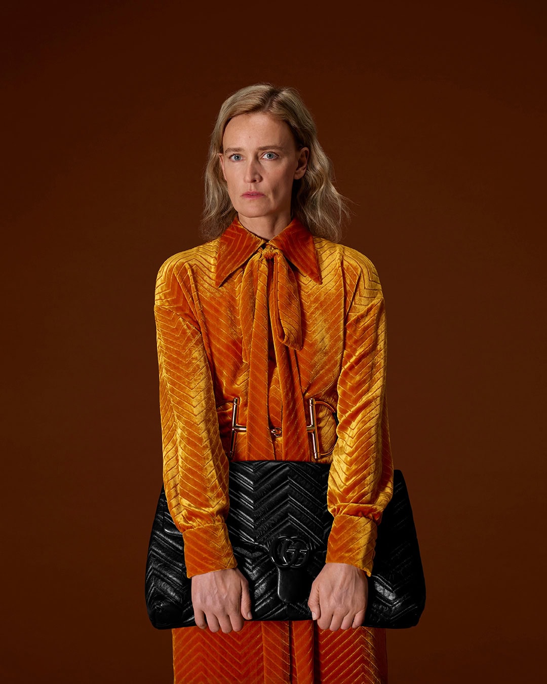 Woman in an orange velvet pussy-bow blouse holding a large quilted black handbag against a warm brown background