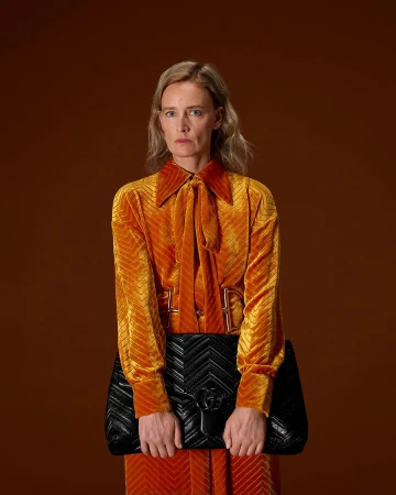 Woman in an orange velvet pussy-bow blouse holding a large quilted black handbag against a warm brown background