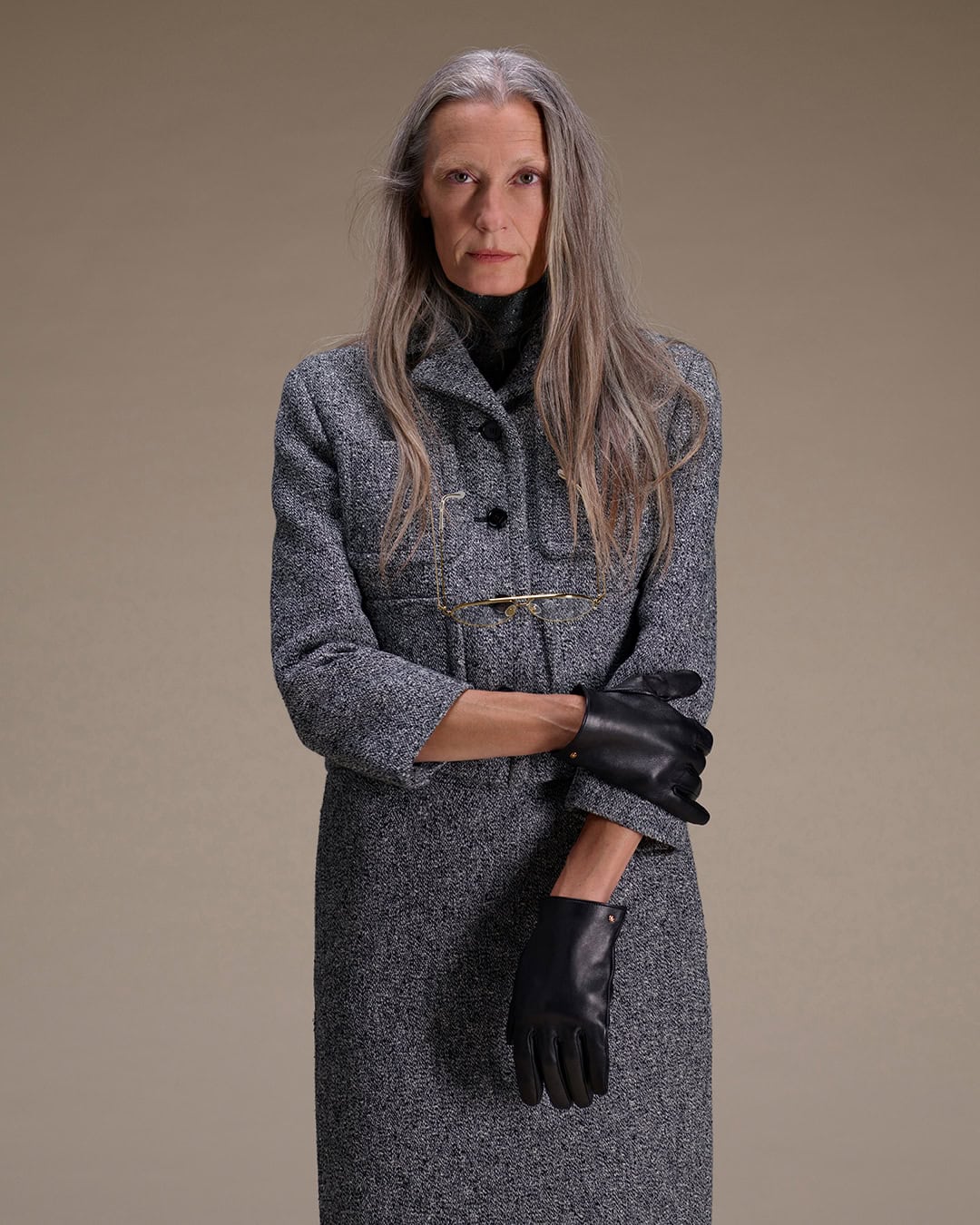 Elderly woman with long grey hair wearing a grey tweed suit and black leather gloves against a neutral background.