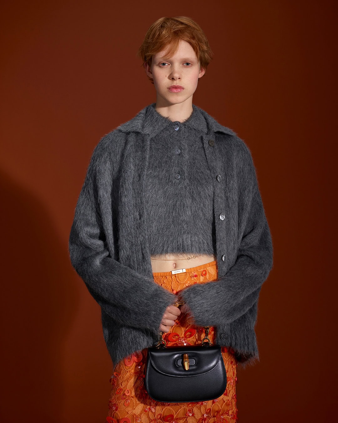 Model with red hair wearing a fuzzy charcoal cardigan over orange skirt holding a black handbag