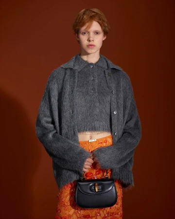 Model with red hair wearing a fuzzy charcoal cardigan over orange skirt holding a black handbag